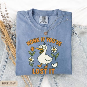 May include: Blue jean-colored t-shirt with the phrase "HONK IF YOU'VE LOST IT" and a cartoon duck illustration. The shirt features a crew neck and short sleeves. A silver necklace with a pendant is draped over the shirt.