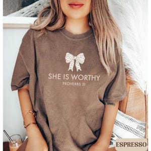 May include: Espresso-colored t-shirt with the text "SHE IS WORTHY" and "PROVERBS 31" printed in a light color. A bow graphic is above the text. The shirt is a short-sleeved, casual style.