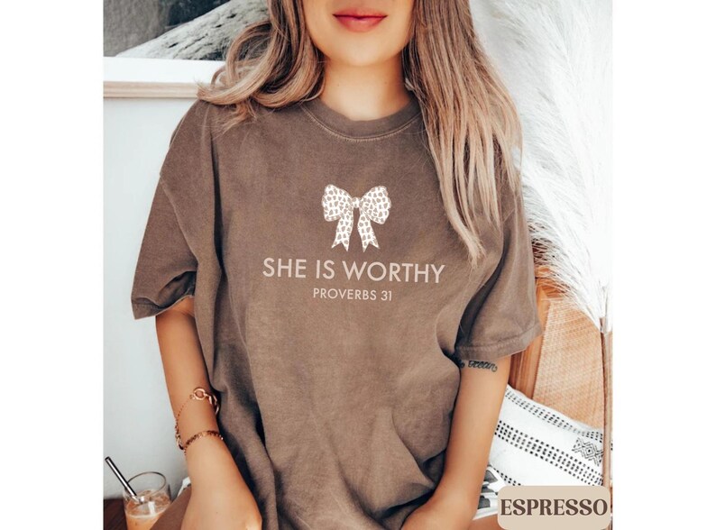 May include: Espresso-colored t-shirt with the text "SHE IS WORTHY" and "PROVERBS 31" printed in a light color. A bow graphic is above the text. The shirt is a short-sleeved, casual style.