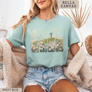 May include: Dusty blue t-shirt with a floral and cross graphic. The graphic features a cross on a hill with flowers in the foreground. The shirt is made of a soft material and has short sleeves. The text "BELLA CANVAS" is printed on the top right.