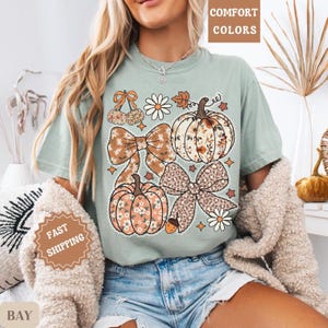 May include: A pale green t-shirt with a fall-themed design of pumpkins, bows, and floral details. The design includes the text "COMFORT COLORS". The shirt has a relaxed fit.