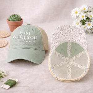 May include: Two baseball caps. One is sage green with a tan mesh back and the text "I AM WITH YOU always". The other cap is turned inside out, showing a dotted pattern on the inside. The caps are on a light-colored surface.