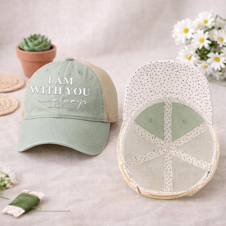 May include: Two baseball caps. One is sage green with a tan mesh back and the text "I AM WITH YOU always". The other cap is turned inside out, showing a dotted pattern on the inside. The caps are on a light-colored surface.