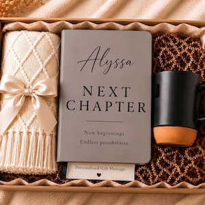 May include: A gift box containing a cream-coloured knit blanket with a satin bow, a grey notebook with the text Alyssa NEXT CHAPTER New beginnings. Endless possibilities., and a black mug with a tan base, all nestled in brown crinkle paper.