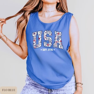 K&ouml;nnte beinhalten: Ein blaues Tanktop mit dem Wort "USA" in einem floralen Muster. Der Text "EST. 1776" befindet sich unter dem Wort "USA". Das Tanktop hat eine einfarbige blaue Farbe.