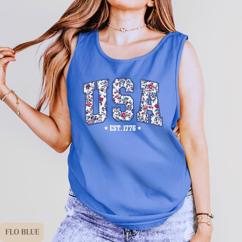 K&ouml;nnte beinhalten: Ein blaues Tanktop mit dem Wort "USA" in einem floralen Muster. Der Text "EST. 1776" befindet sich unter dem Wort "USA". Das Tanktop hat eine einfarbige blaue Farbe.