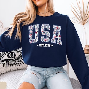 Peut inclure: Sweat-shirt bleu marine avec le mot "USA" en motif floral sur la poitrine. Le texte "EST. 1776" est en dessous. Le sweat-shirt est &agrave; col rond et &agrave; manches longues.