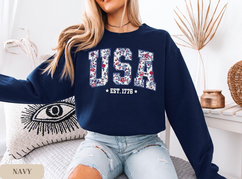Peut inclure: Sweat-shirt bleu marine avec le mot "USA" en motif floral sur la poitrine. Le texte "EST. 1776" est en dessous. Le sweat-shirt est &agrave; col rond et &agrave; manches longues.