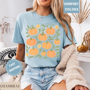 May include: Light blue t-shirt featuring a pumpkin design. The design includes orange pumpkins, green vines, and yellow flowers. The words "COMFORT COLORS" are printed on the top right of the shirt.
