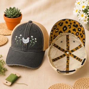 May include: A dark gray trucker hat with a tan mesh back and a white chicken embroidered on the front. The hat's interior features a leopard print design. A second hat is shown with the interior facing up.