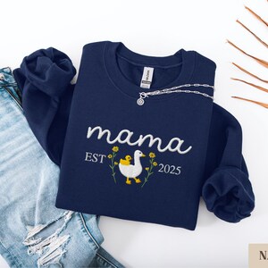 May include: Navy blue sweatshirt with embroidered text "mama est 2025" and a white duck with a yellow chick. The duck is surrounded by yellow flowers.