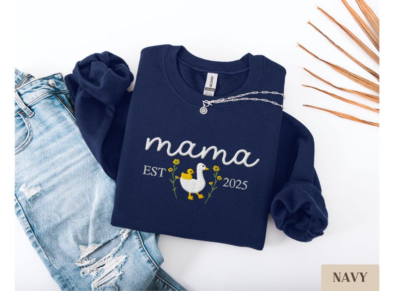 May include: Navy blue sweatshirt with embroidered text "mama est 2025" and a white duck with a yellow chick. The duck is surrounded by yellow flowers.