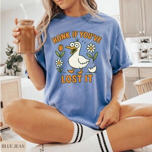May include: A blue t-shirt with the phrase "HONK IF YOU'VE LOST IT" and a cartoon duck graphic. The duck is white with an orange beak and feet, surrounded by flowers and an eggshell. The shirt is worn by a person.