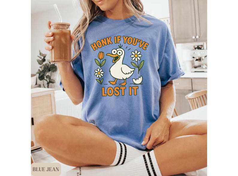 May include: A blue t-shirt with the phrase "HONK IF YOU'VE LOST IT" and a cartoon duck graphic. The duck is white with an orange beak and feet, surrounded by flowers and an eggshell. The shirt is worn by a person.