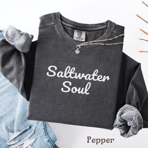 Op de afbeelding: Een donkergrijs sweatshirt met lange mouwen met de geborduurde tekst "Saltwater Soul" in het wit. De sweatshirt heeft opgerolde mouwen en een zilveren ketting. Het woord "Pepper" staat onderaan gedrukt.