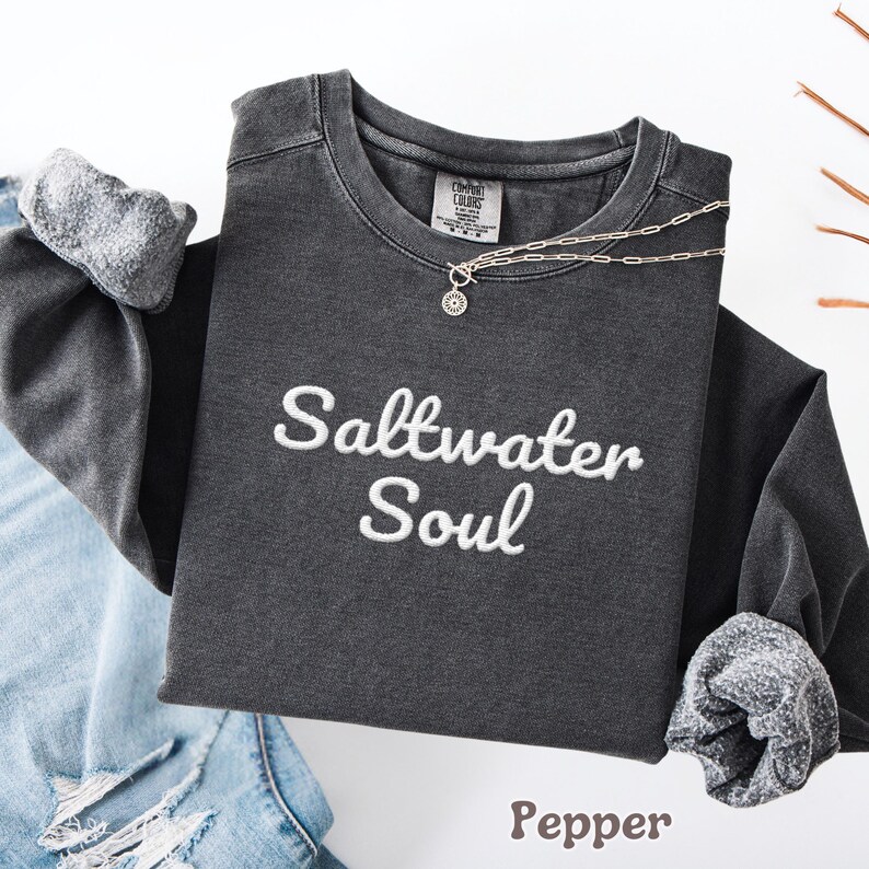 Op de afbeelding: Een donkergrijs sweatshirt met lange mouwen met de geborduurde tekst "Saltwater Soul" in het wit. De sweatshirt heeft opgerolde mouwen en een zilveren ketting. Het woord "Pepper" staat onderaan gedrukt.
