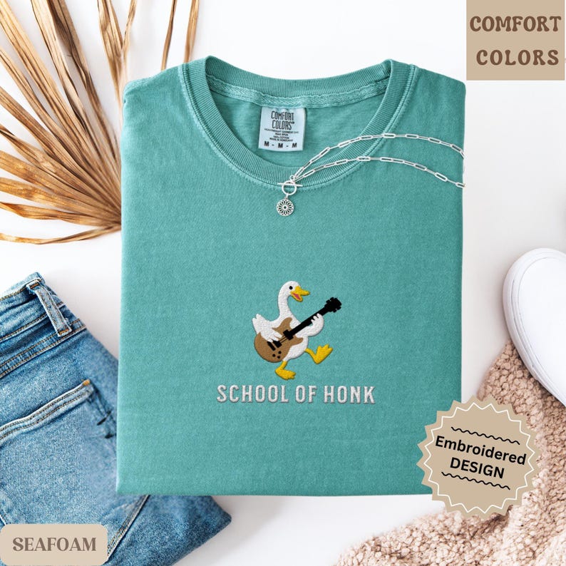 May include: Seafoam green t-shirt with an embroidered design of a goose playing a guitar, with the text "SCHOOL OF HONK" below. A silver necklace with a pendant is on the shirt. The shirt is folded and placed on a white surface with a pair of blue jeans.