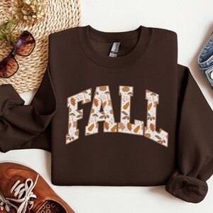 K&ouml;nnte beinhalten: Braunes Sweatshirt mit einem wei&szlig;en "FALL"-Motiv. Die Buchstaben sind mit einem Muster aus Herbstbl&auml;ttern, Eicheln und K&uuml;rbissen gef&uuml;llt.