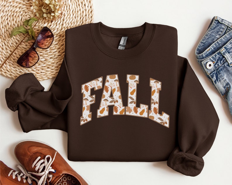 K&ouml;nnte beinhalten: Braunes Sweatshirt mit einem wei&szlig;en "FALL"-Motiv. Die Buchstaben sind mit einem Muster aus Herbstbl&auml;ttern, Eicheln und K&uuml;rbissen gef&uuml;llt.