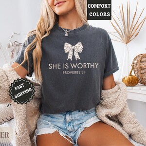 May include: Dark gray t-shirt with the text "SHE IS WORTHY" and a bow design. The shirt also includes the text "PROVERBS 31". The model is wearing denim shorts and a cream-colored cardigan.