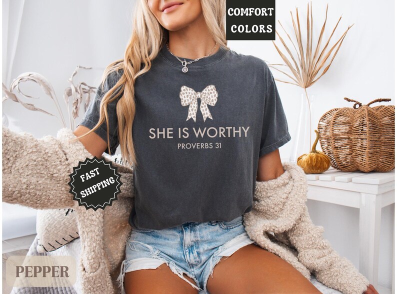 May include: Dark gray t-shirt with the text "SHE IS WORTHY" and a bow design. The shirt also includes the text "PROVERBS 31". The model is wearing denim shorts and a cream-colored cardigan.