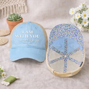 May include: Two baseball caps. One is light blue with the text "I AM WITH YOU always" in white. The other cap is turned inside out, revealing a floral pattern lining. Both caps have a tan mesh back.