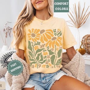 May include: A butter yellow short-sleeve t-shirt featuring a large graphic print of stylized sunflowers and foliage in shades of mustard yellow, sage green, and cream. The design has a distressed texture and the shirt is worn with denim shorts.