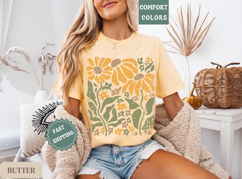 May include: A butter yellow short-sleeve t-shirt featuring a large graphic print of stylized sunflowers and foliage in shades of mustard yellow, sage green, and cream. The design has a distressed texture and the shirt is worn with denim shorts.