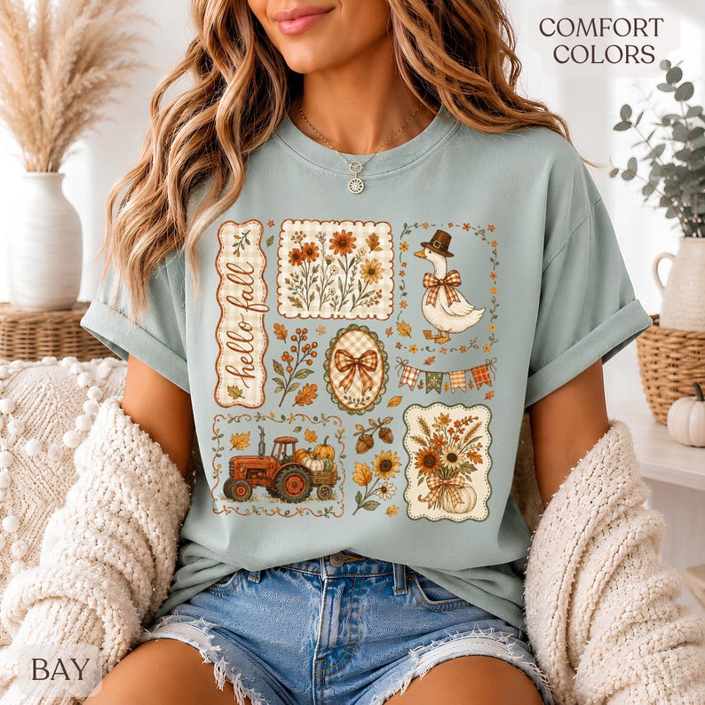 May include: A light blue t-shirt featuring a graphic print with the text "hello fall" and various autumn-themed illustrations including a tractor, a goose wearing a hat, sunflowers, pumpkins, and plaid patterns arranged in a collage style.