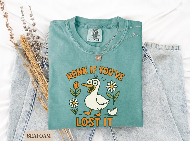 May include: Seafoam green t-shirt with the phrase "HONK IF YOU'VE LOST IT" and a cartoon duck illustration. The shirt is folded on top of a pair of blue jeans. A silver necklace with a pendant is on the shirt.