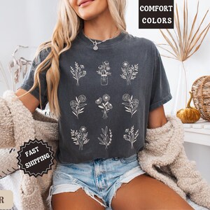 May include: A dark gray t-shirt with a white floral design. The shirt has the text "Comfort Colors" printed on the top and "Fast Shipping" printed on the bottom. The model is wearing the shirt with denim shorts.