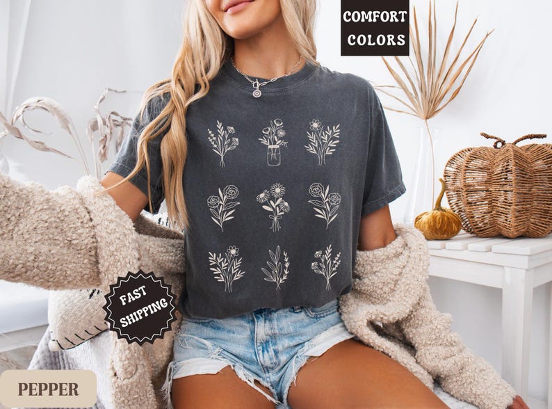 May include: A dark gray t-shirt with a white floral design. The shirt has the text "Comfort Colors" printed on the top and "Fast Shipping" printed on the bottom. The model is wearing the shirt with denim shorts.