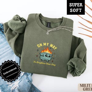 Pu&ograve; includere: Felpa verde militare con la scritta ricamata "ON MY WAY To Brighten Your Day" e una grafica di un bidone della spazzatura sorridente in fiamme. La felpa &egrave; piegata accanto a un paio di jeans.