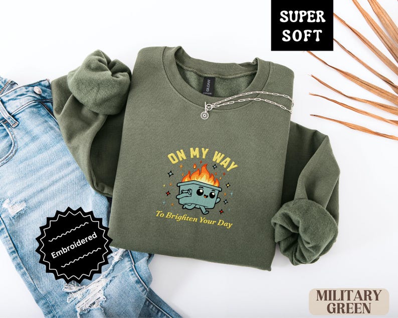 Pu&ograve; includere: Felpa verde militare con la scritta ricamata "ON MY WAY To Brighten Your Day" e una grafica di un bidone della spazzatura sorridente in fiamme. La felpa &egrave; piegata accanto a un paio di jeans.