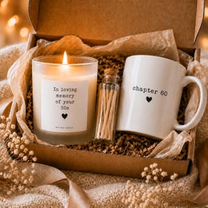May include: A gift box containing a lit soy wax candle with a label reading In loving memory of your 50s with a heart icon, a small glass bottle filled with wooden matches, and a white ceramic mug with the text chapter 60 and a heart icon.
