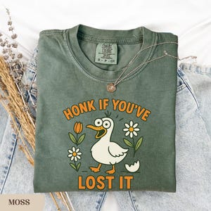May include: Moss green t-shirt with a humorous graphic of a cartoon duck with the text "HONK IF YOU'VE LOST IT". The design includes orange tulips, white daisies, and a cracked egg. A silver necklace rests on the shirt.