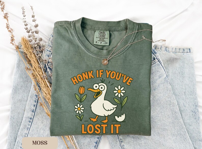 May include: Moss green t-shirt with a humorous graphic of a cartoon duck with the text "HONK IF YOU'VE LOST IT". The design includes orange tulips, white daisies, and a cracked egg. A silver necklace rests on the shirt.