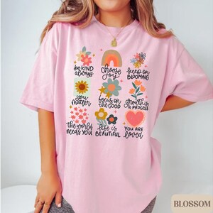 Pu&ograve; includere: Maglietta rosa con un design grafico di fiori colorati e testo che dice "be kind always", "choose joy", "keep on blooming", "you matter", "focus on the good", "growth is a process", "the world needs you", "life is beautiful" e "you are loved".
