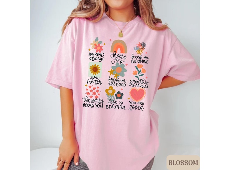 Pu&ograve; includere: Maglietta rosa con un design grafico di fiori colorati e testo che dice "be kind always", "choose joy", "keep on blooming", "you matter", "focus on the good", "growth is a process", "the world needs you", "life is beautiful" e "you are loved".
