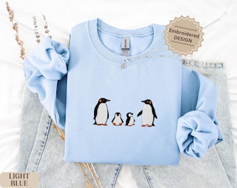Peut inclure: Sweat-shirt bleu clair avec une broderie d'une famille de pingouins. Les pingouins sont noirs et blancs avec des détails orange. Le texte "Embroidered DESIGN" est dans une forme d'étoile. Le texte "LIGHT BLUE" est en bas.