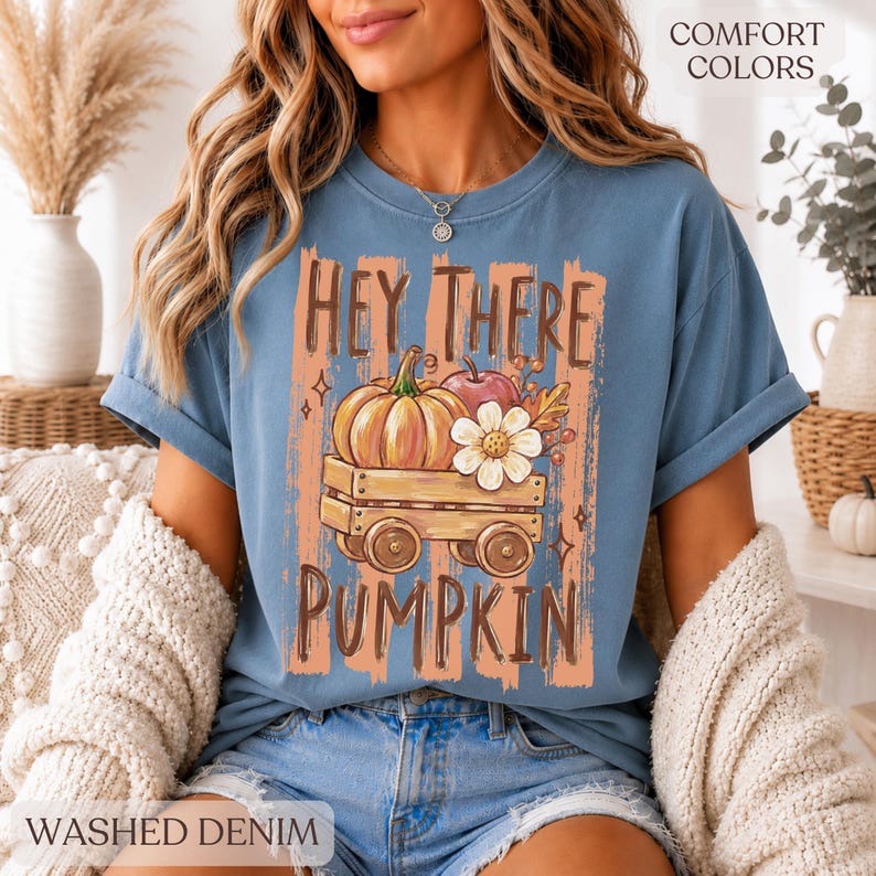 Op de afbeelding: Een blauw t-shirt met korte mouwen en een afbeelding van een houten wagen met een pompoen, een appel en een witte bloem. De tekst "HEY THERE PUMPKIN" staat in bruine letters boven en onder de illustratie van de wagen op een perzikkleurige achtergrond.