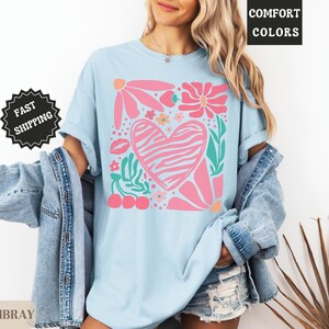 May include: Light blue t-shirt with a pink zebra-striped heart design, surrounded by pink flowers, green leaves, a strawberry, cherries, and a kiss. The shirt has the words "Comfort Colors" and "Chambray" printed on it, and a "Fast Shipping" sticker.