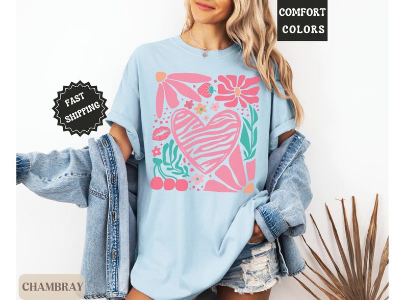 May include: Light blue t-shirt with a pink zebra-striped heart design, surrounded by pink flowers, green leaves, a strawberry, cherries, and a kiss. The shirt has the words "Comfort Colors" and "Chambray" printed on it, and a "Fast Shipping" sticker.
