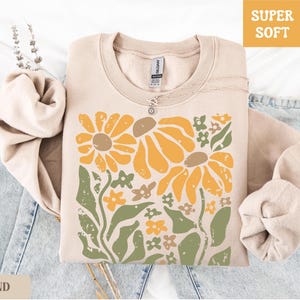 May include: A sand-colored sweatshirt with a floral design featuring large yellow flowers, green leaves, and small orange and brown flowers. The words "SUPER SOFT" are in a yellow box. A silver necklace is on the sweatshirt.