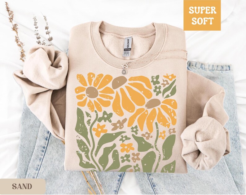 May include: A sand-colored sweatshirt with a floral design featuring large yellow flowers, green leaves, and small orange and brown flowers. The words "SUPER SOFT" are in a yellow box. A silver necklace is on the sweatshirt.
