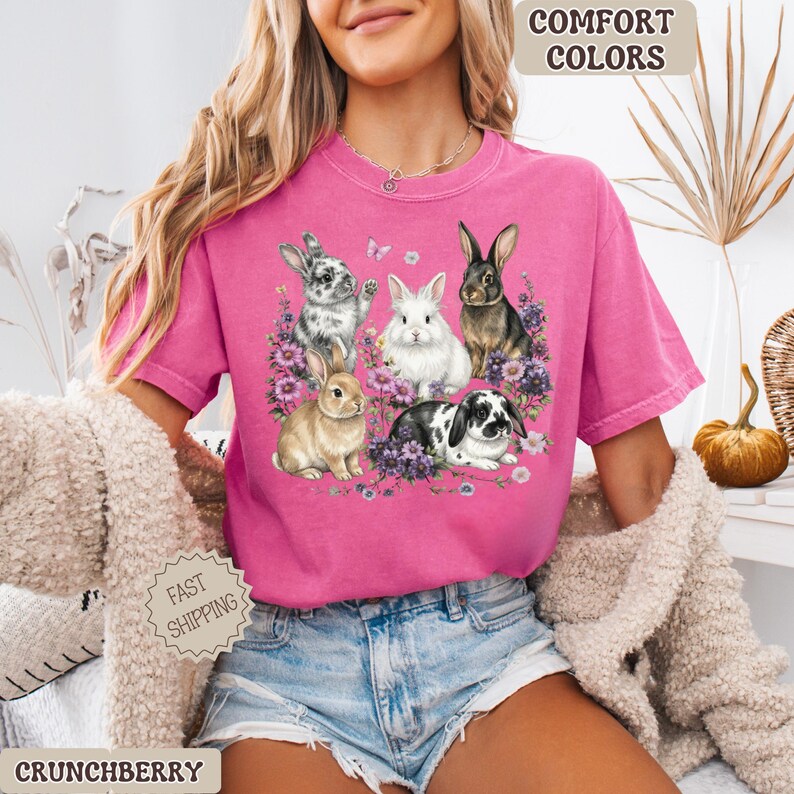 Puede incluir: Una camiseta rosa con un gr&aacute;fico de cinco conejos rodeados de flores moradas y mariposas. La camiseta tiene las palabras "Comfort Colors" impresas en la parte superior derecha y "Fast Shipping" en la parte inferior izquierda.