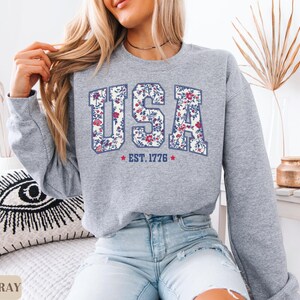 Peut inclure: Un sweat-shirt gris clair avec le mot "USA" dans un motif floral, avec "EST. 1776" en dessous. Le sweat-shirt est de style col rond et est associ&eacute; &agrave; un jean bleu. Les mots "SPORT GRAY" sont au bas de l'image.