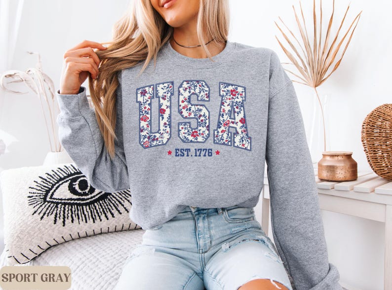 Peut inclure: Un sweat-shirt gris clair avec le mot "USA" dans un motif floral, avec "EST. 1776" en dessous. Le sweat-shirt est de style col rond et est associ&eacute; &agrave; un jean bleu. Les mots "SPORT GRAY" sont au bas de l'image.