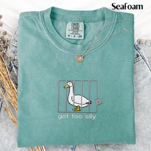 May include: Seafoam green Comfort Colors t-shirt with an embroidered duck in a cage design. The text "got too silly" is below the duck. A silver necklace with a pendant is on the shirt. The shirt is folded on top of denim jeans.