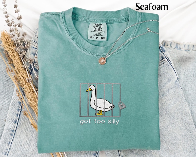 May include: Seafoam green Comfort Colors t-shirt with an embroidered duck in a cage design. The text "got too silly" is below the duck. A silver necklace with a pendant is on the shirt. The shirt is folded on top of denim jeans.